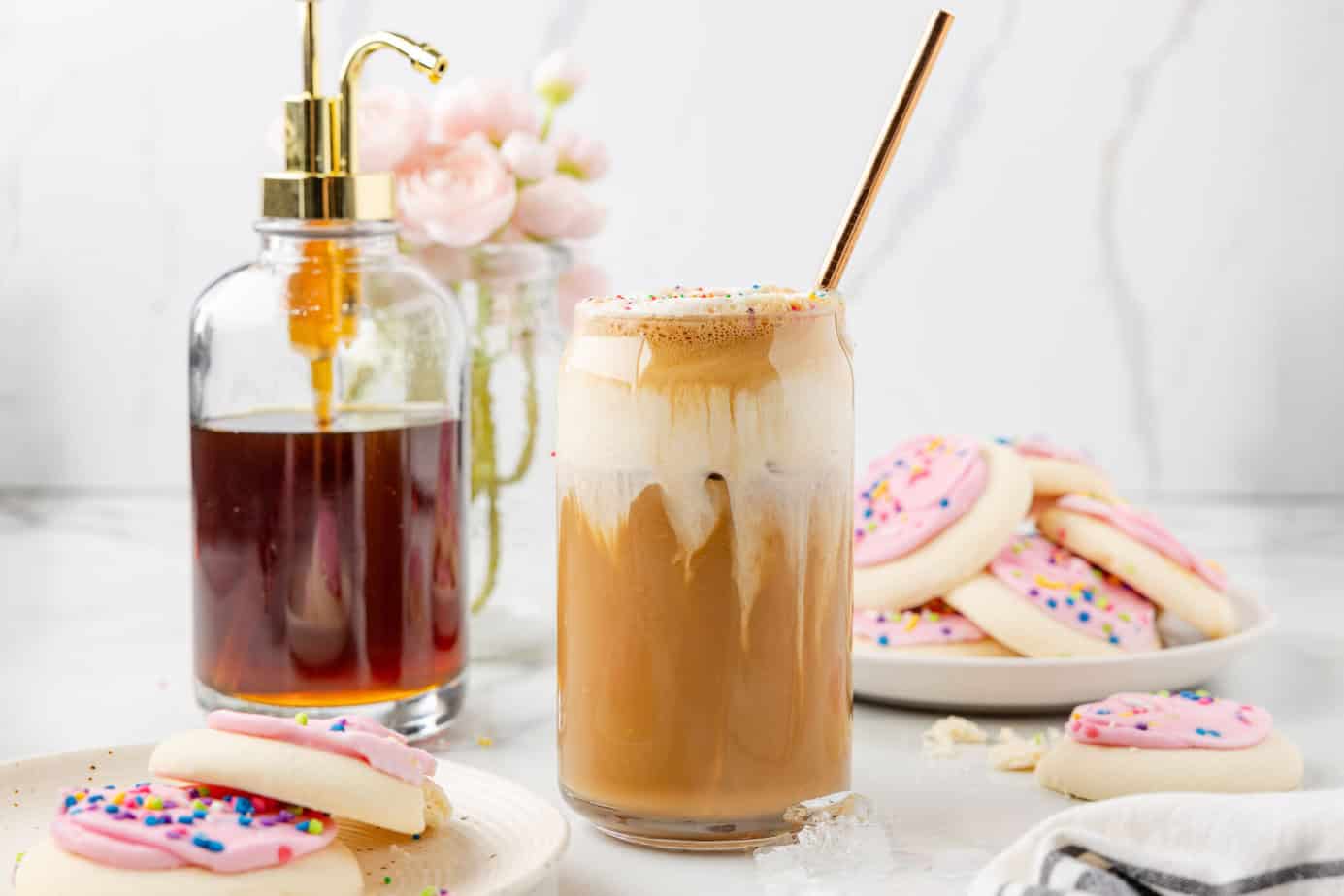 Sugar Cookie Latte with the almond milk foam seeping into the espresso layer. Pink sugar cookies and sugar cookie syrup are in the background.