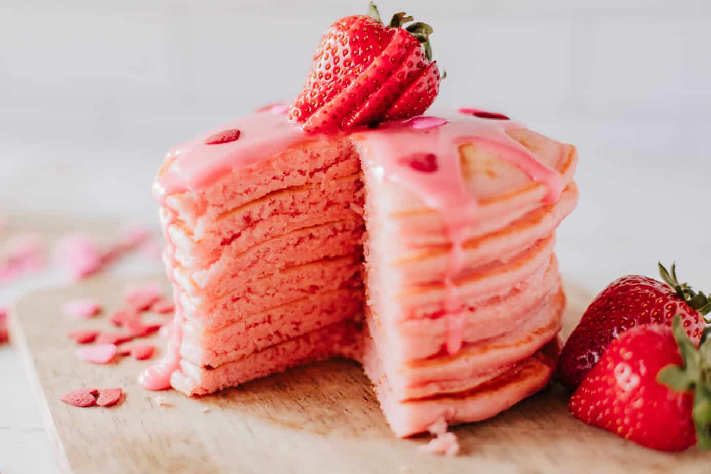 strawberry pancakes stacked on a cutting board with pink syrup, heart sprinkles, and fresh strawberries
