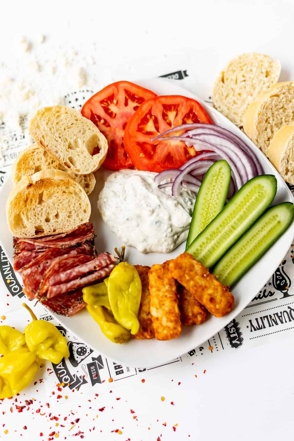 bread, tomatoes, pickles, onion, hashbrowns, peppers and salami on a plate around the Tzatziki dip