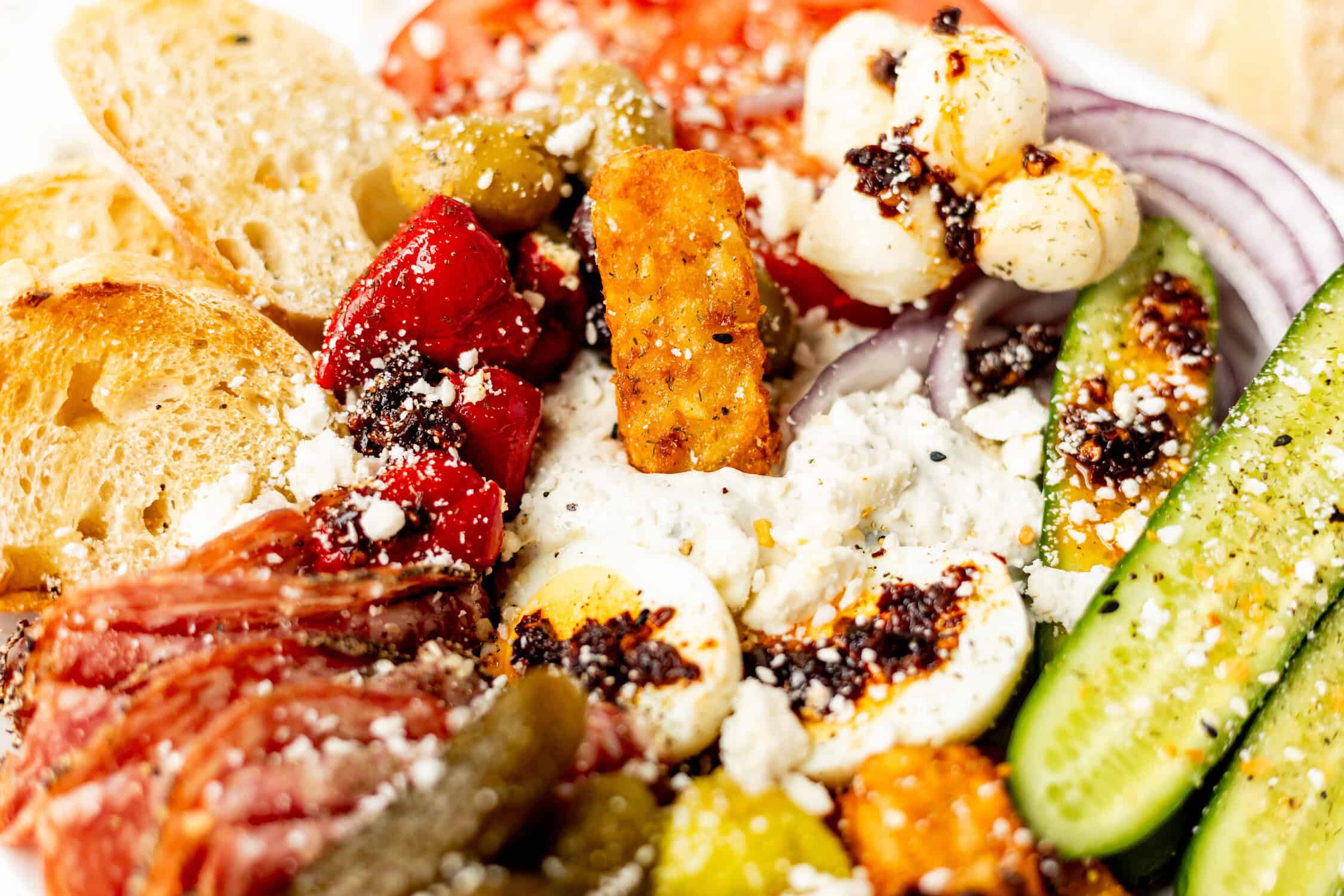 Freshly prepared savory girl snack plate featuring pickles, red onions, tomatoes, kalamata olives, feta cheese, peppered with herbs and olive oil, served with toasted bread slices.