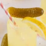 dill pickle lemonade in a glass with pickles and lemons and a pink straw