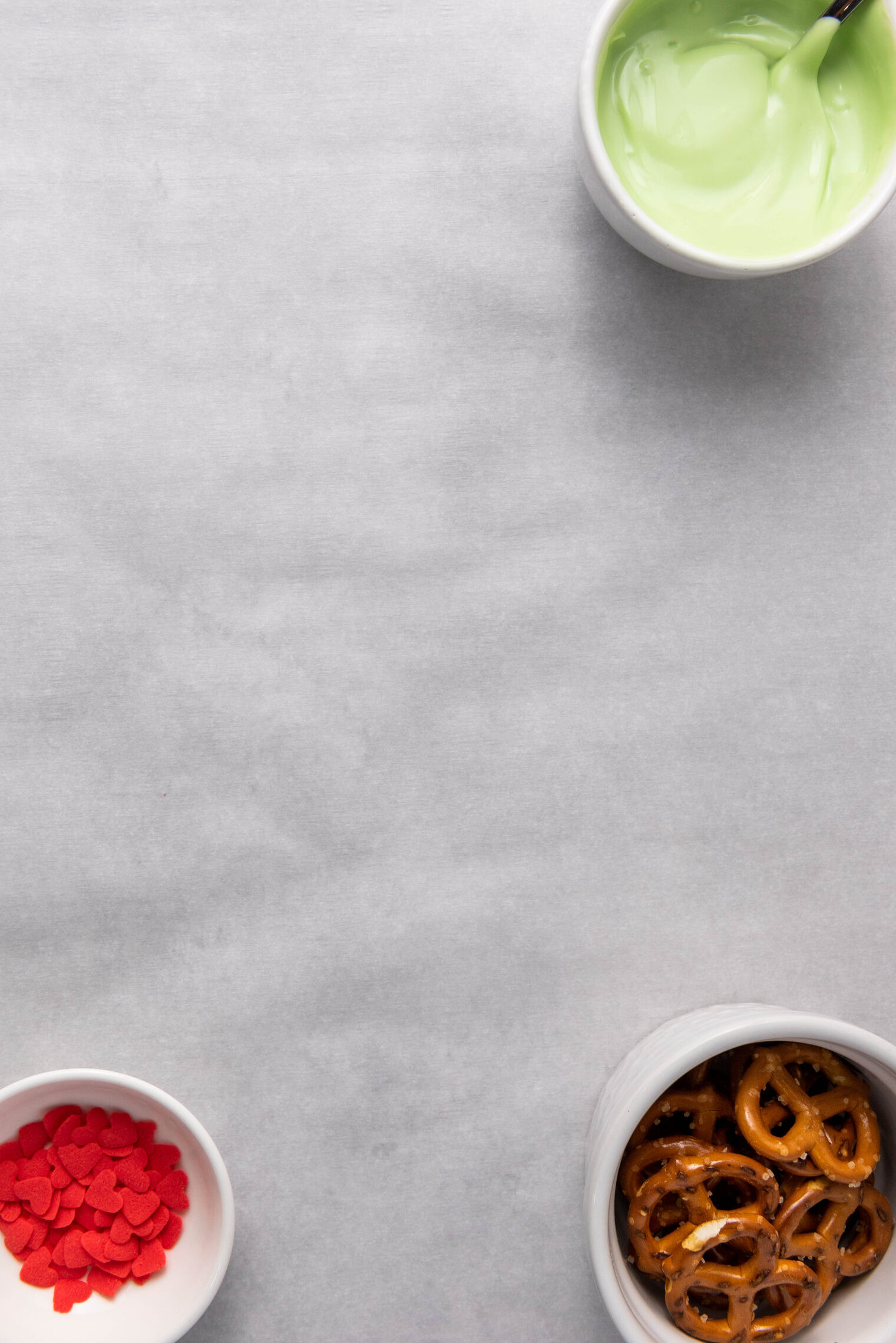 melted green candy melts, heart sprinkles and pretzels in bowls on parchment paper